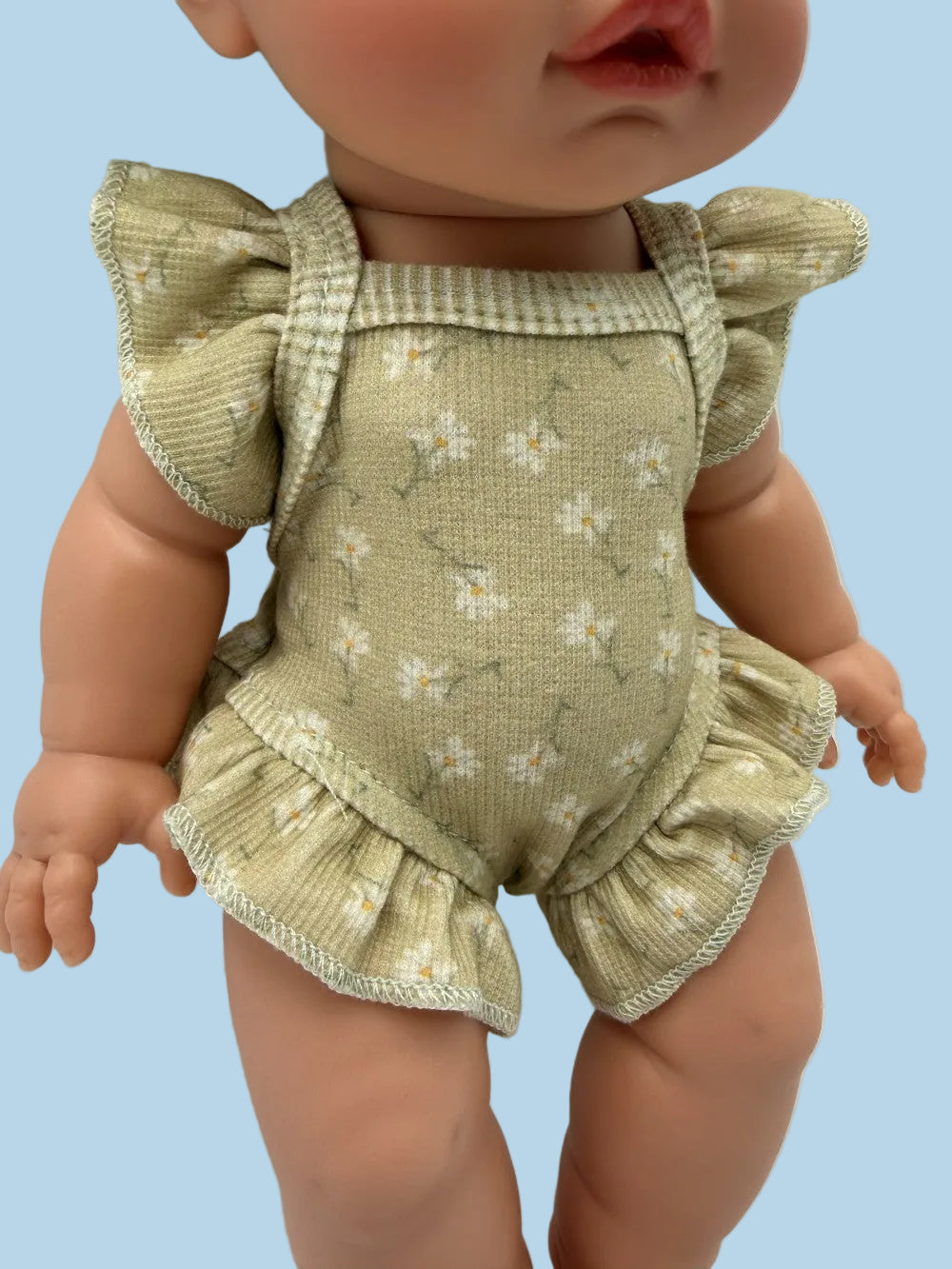 A close-up of a doll wearing a ribbed sage green romper with a small white floral print, ruffled shoulder straps, and ruffled leg openings.