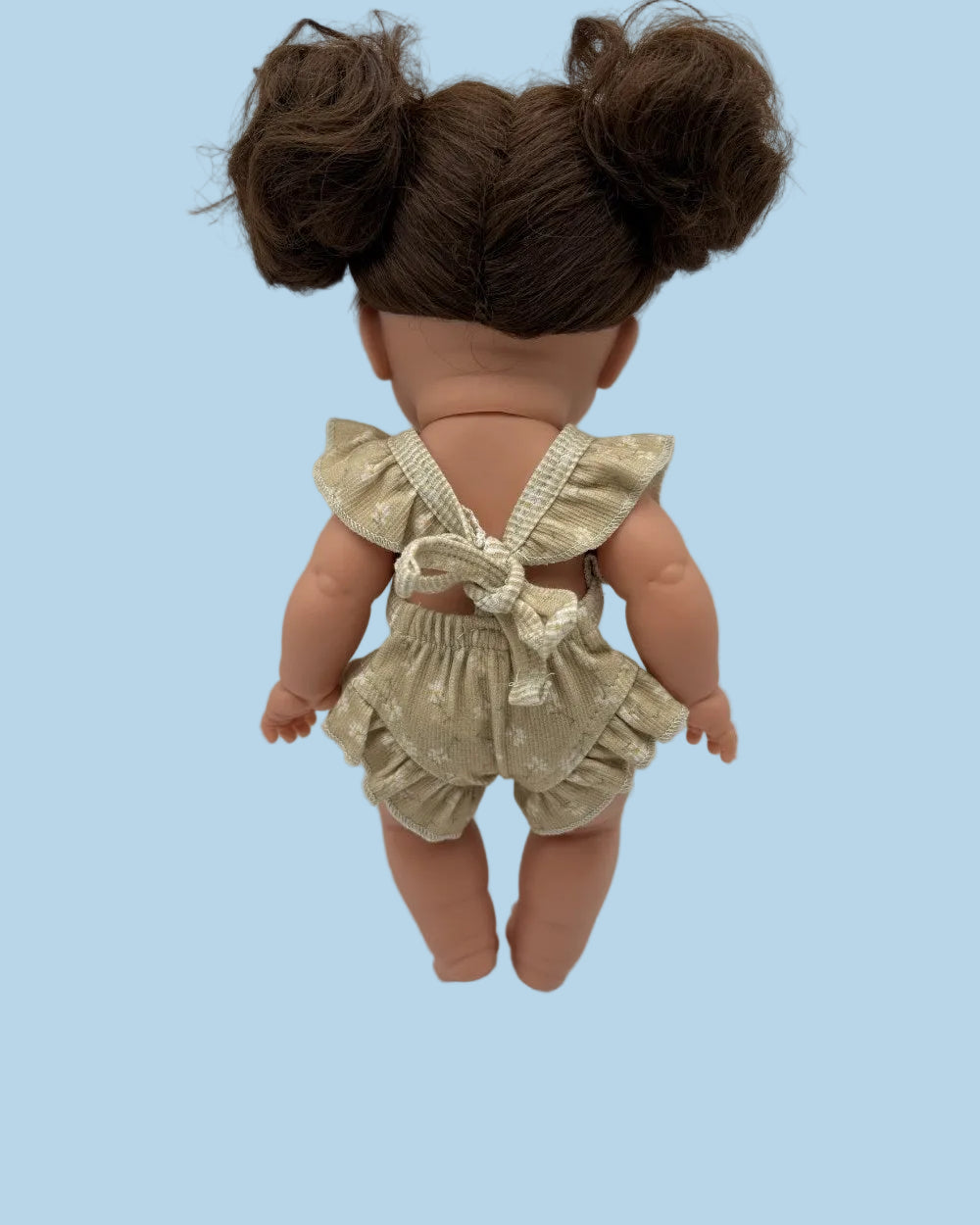 Back view of a doll with brown hair in two buns, wearing a beige and white ruffled romper with a bow on the back, set against a plain light gray background.