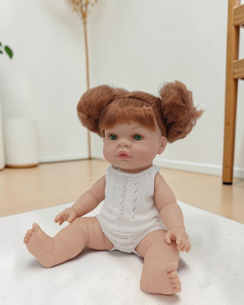 Realistic baby doll with red hair styled in buns sitting on a white surface.