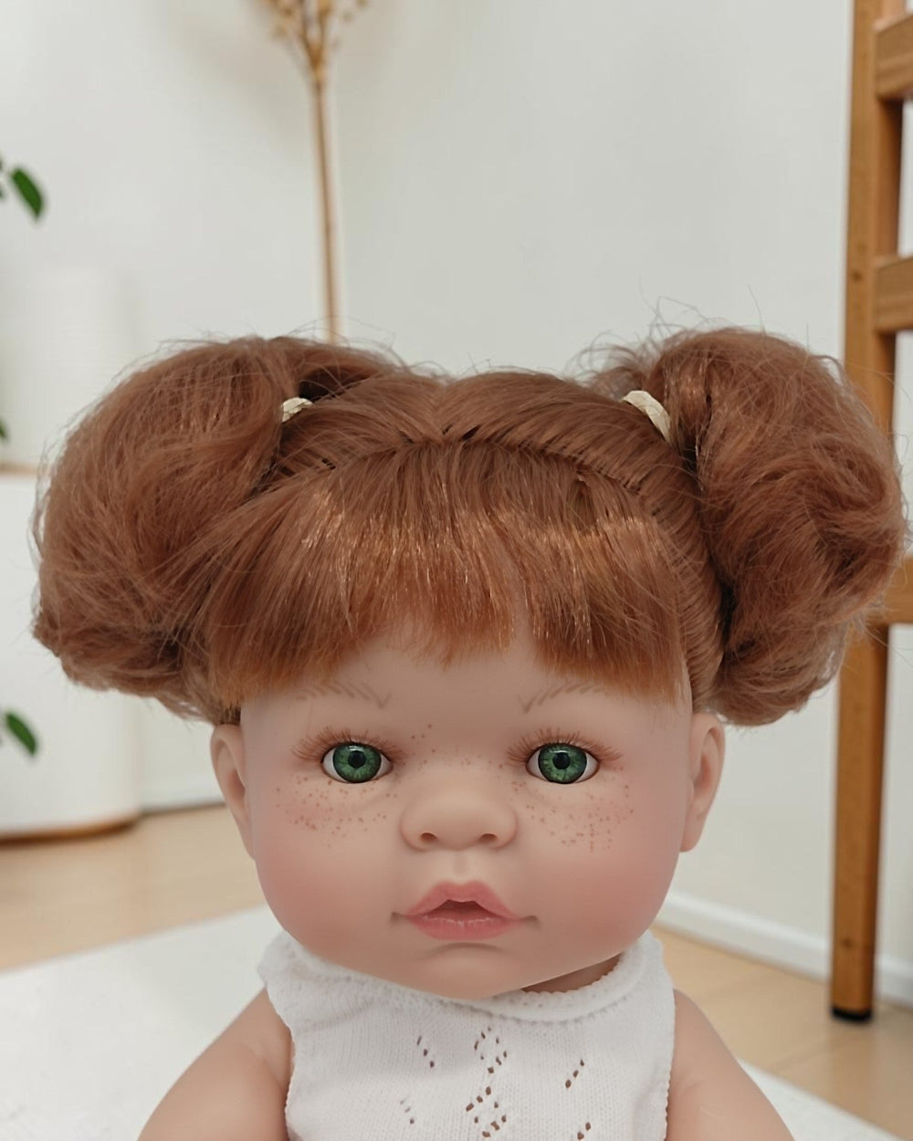 Doll with red hair styled in buns wearing a white outfit indoors.