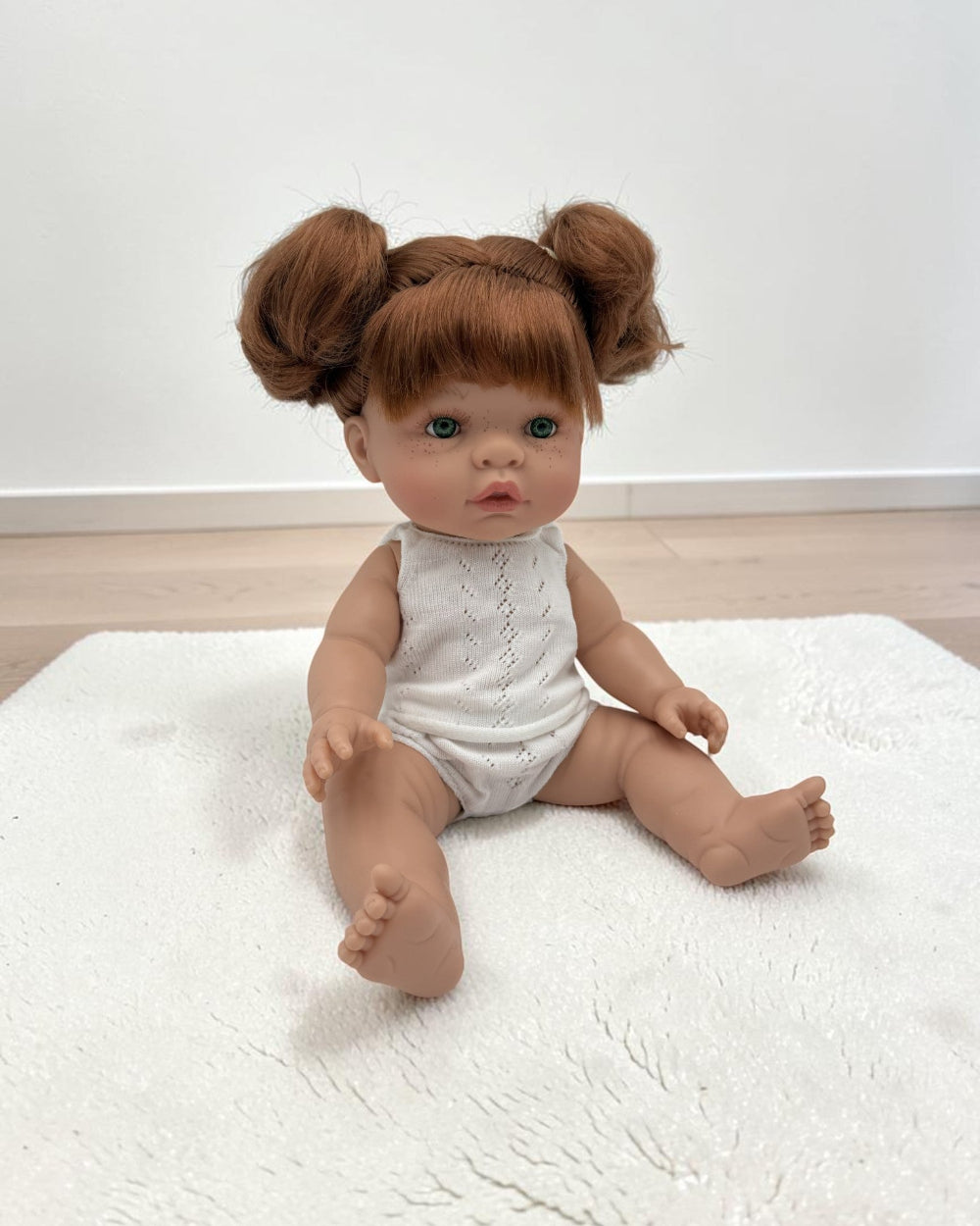 A realistic baby doll with red hair in two buns sits on a white textured rug. The doll has a realistic skin tone, blue eyes, and is wearing a white sleeveless knit romper.