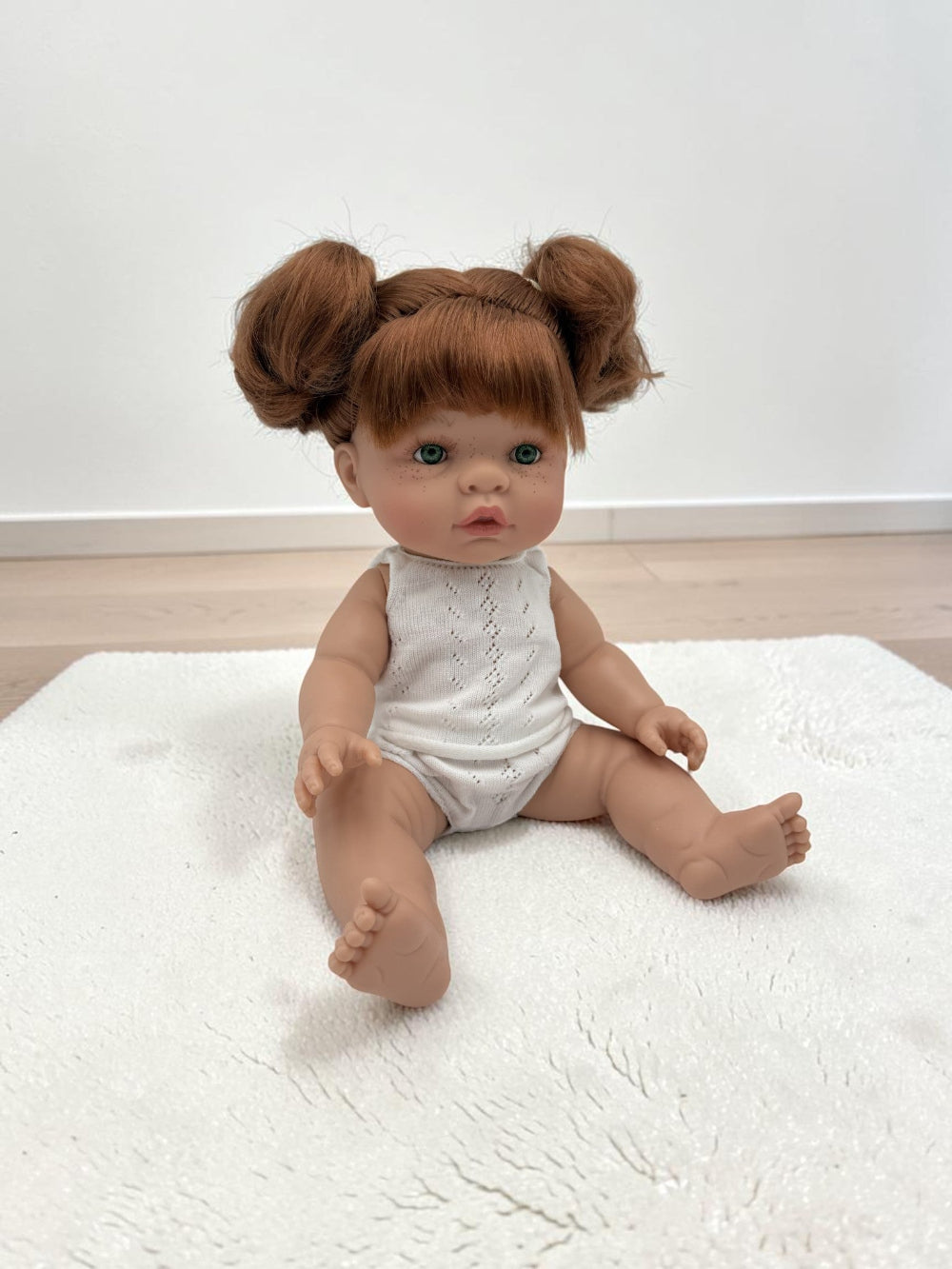 A realistic baby doll with red hair in two buns sits on a white textured rug. The doll has a realistic skin tone, blue eyes, and is wearing a white sleeveless knit romper.