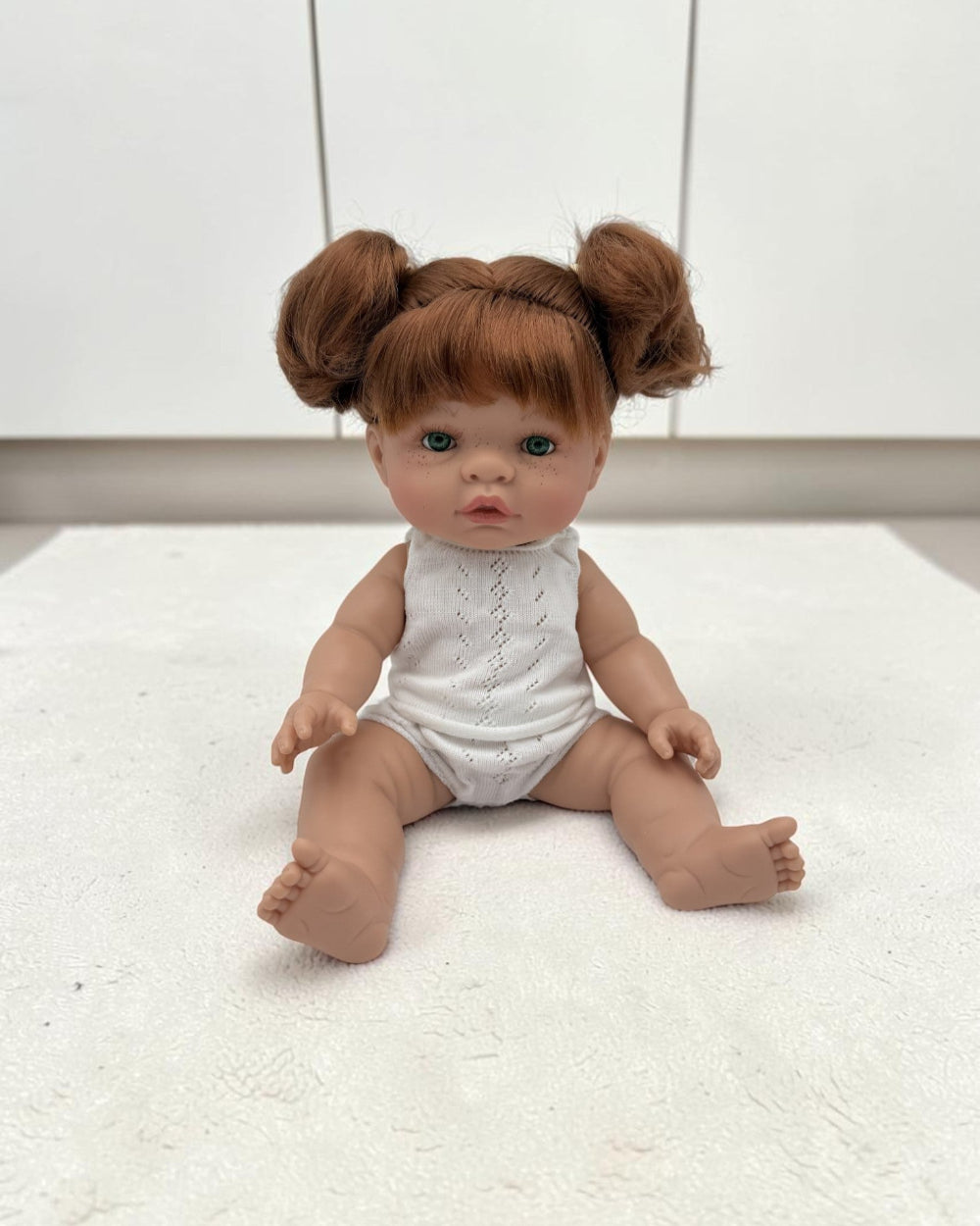 A realistic baby doll with red hair styled in two buns, wearing a white sleeveless top and diaper, sitting on a white textured surface against a white paneled background.