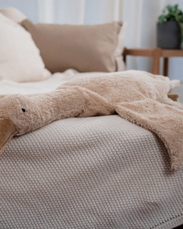 Cream Senger goose cuddly toy laying on knit blanket in modern bedroom setting