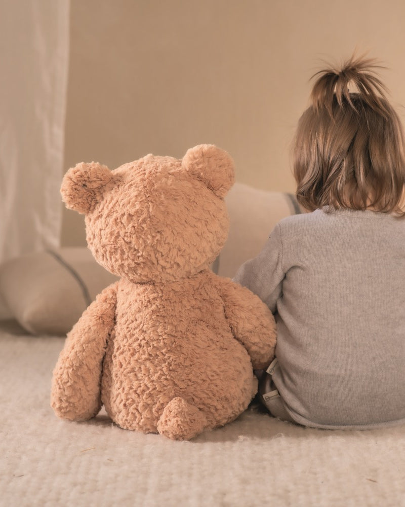 Child sitting on a bed with a large teddy bear, facing away from the camera.