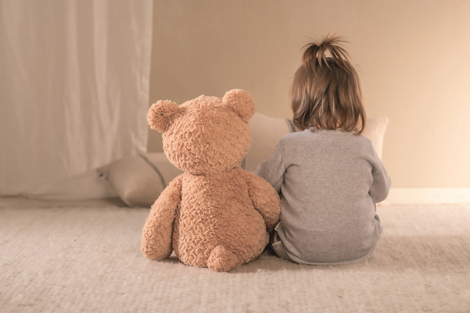 Child sitting on a bed with a large teddy bear, facing away from the camera.