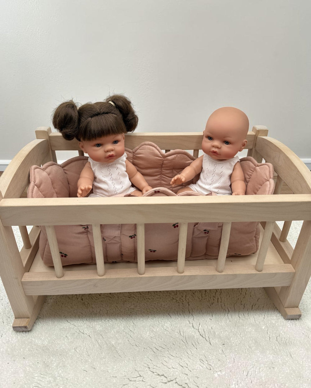 A light natural wood baby cradle with a pink quilted cushion, holding two dolls against a plain light gray wall and white textured floor.