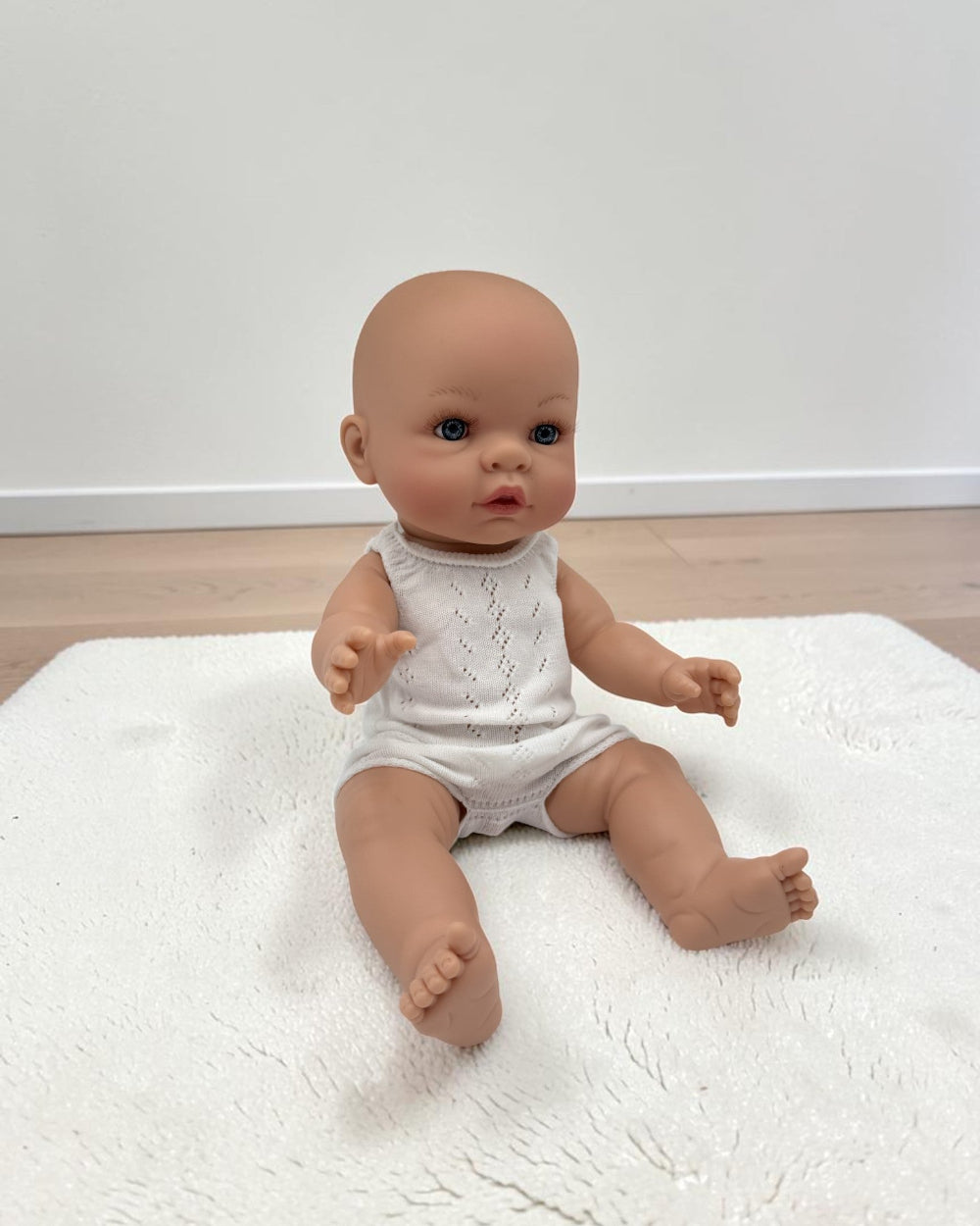 A realistic baby doll with a bald head and blue eyes, wearing a white sleeveless knit romper, sitting on a white textured rug against a plain white wall and light wood floor.