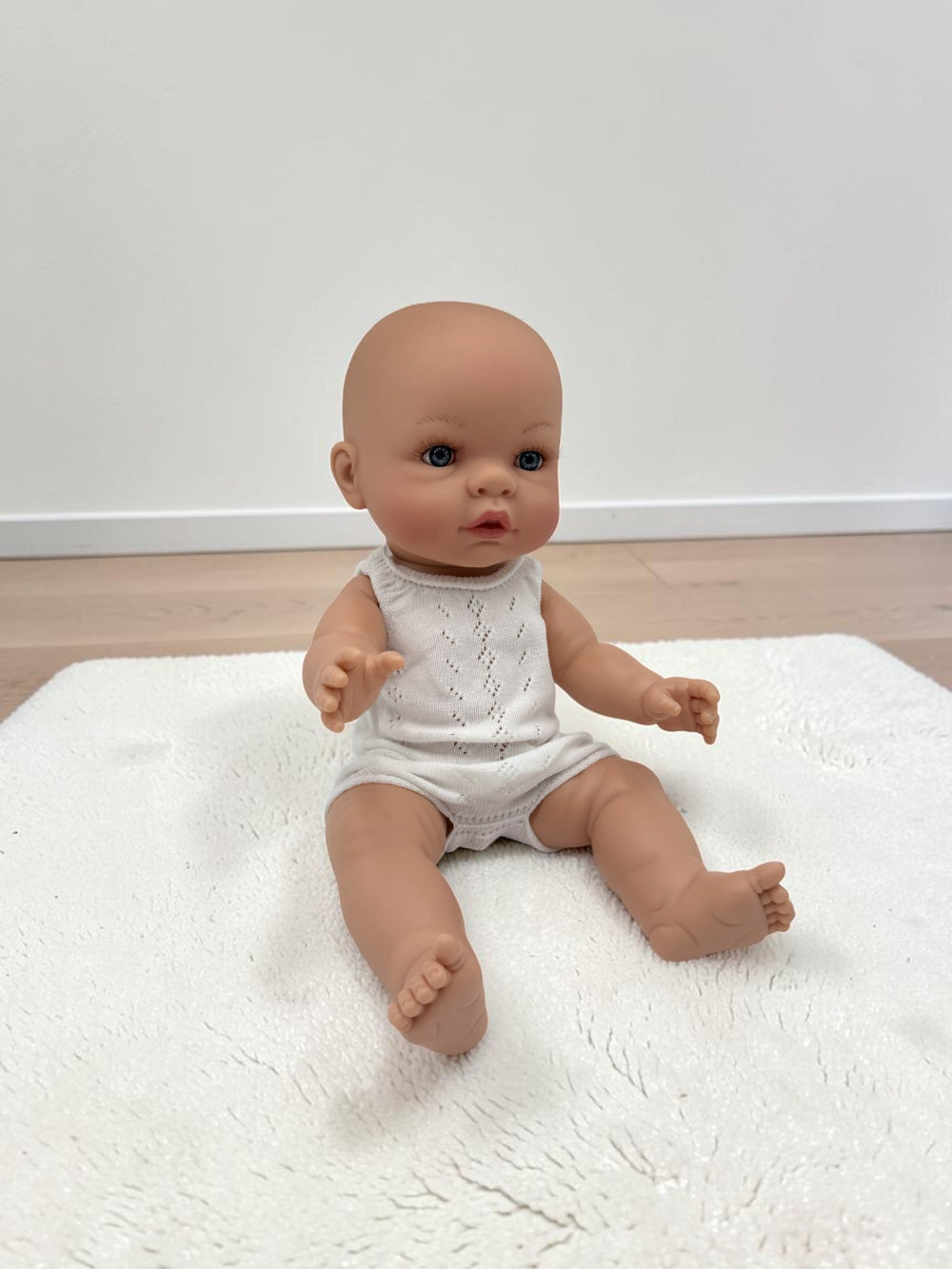 A realistic baby doll with a bald head and blue eyes, wearing a white sleeveless knit romper, sitting on a white textured rug against a plain white wall and light wood floor.