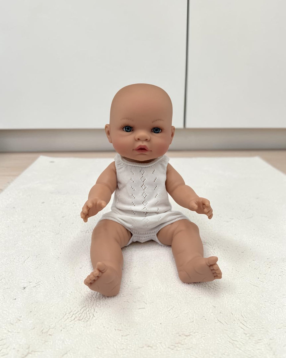 A realistic baby doll with a bald head and blue eyes, wearing a white sleeveless romper, sitting on a white textured rug in front of white cabinets.