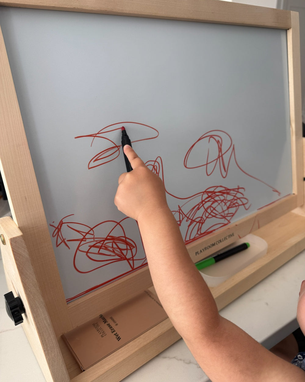 Child drawing on a whiteboard with red marker, holding a black marker.
