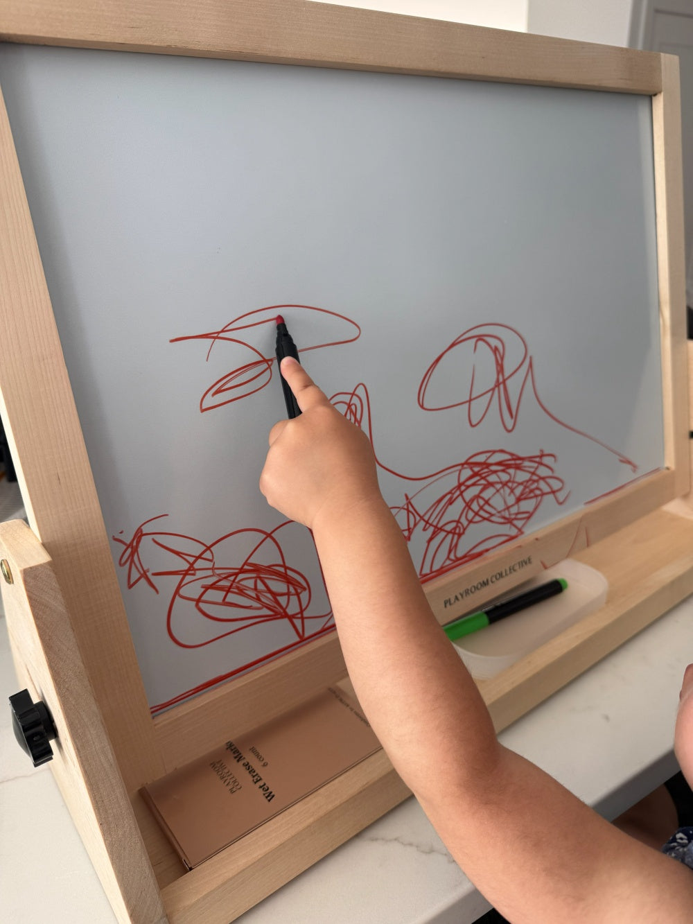Child drawing on a whiteboard with red marker, holding a black marker.