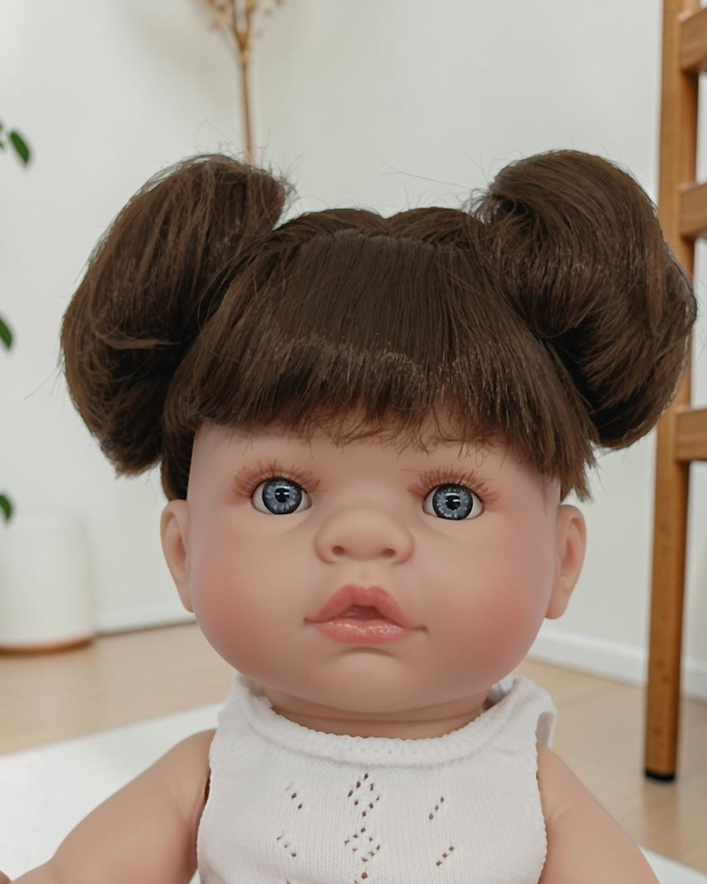 Realistic baby doll with brown hair styled in buns, wearing a white sleeveless top.