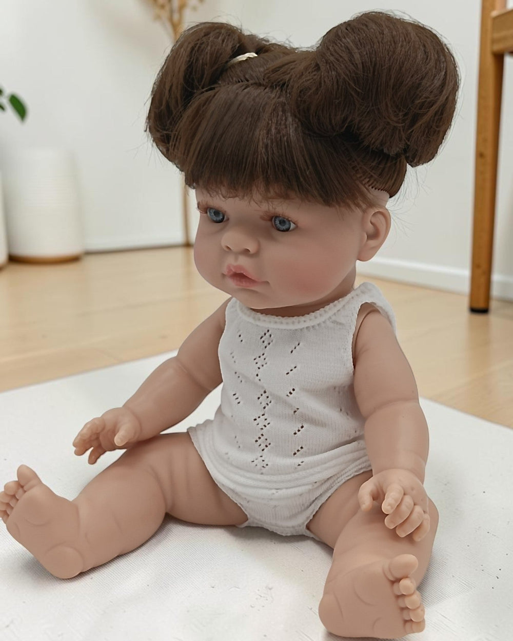 Realistic baby doll with brown hair styled in buns, wearing a white lace romper, on a light-colored surface.