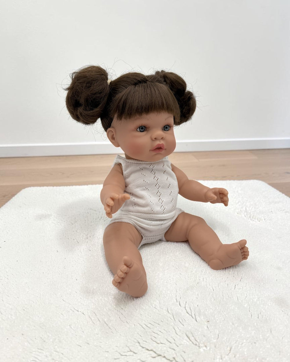 A realistic baby doll with brown hair in two buns sits on a white textured rug in a room with light wood flooring and a white wall.
