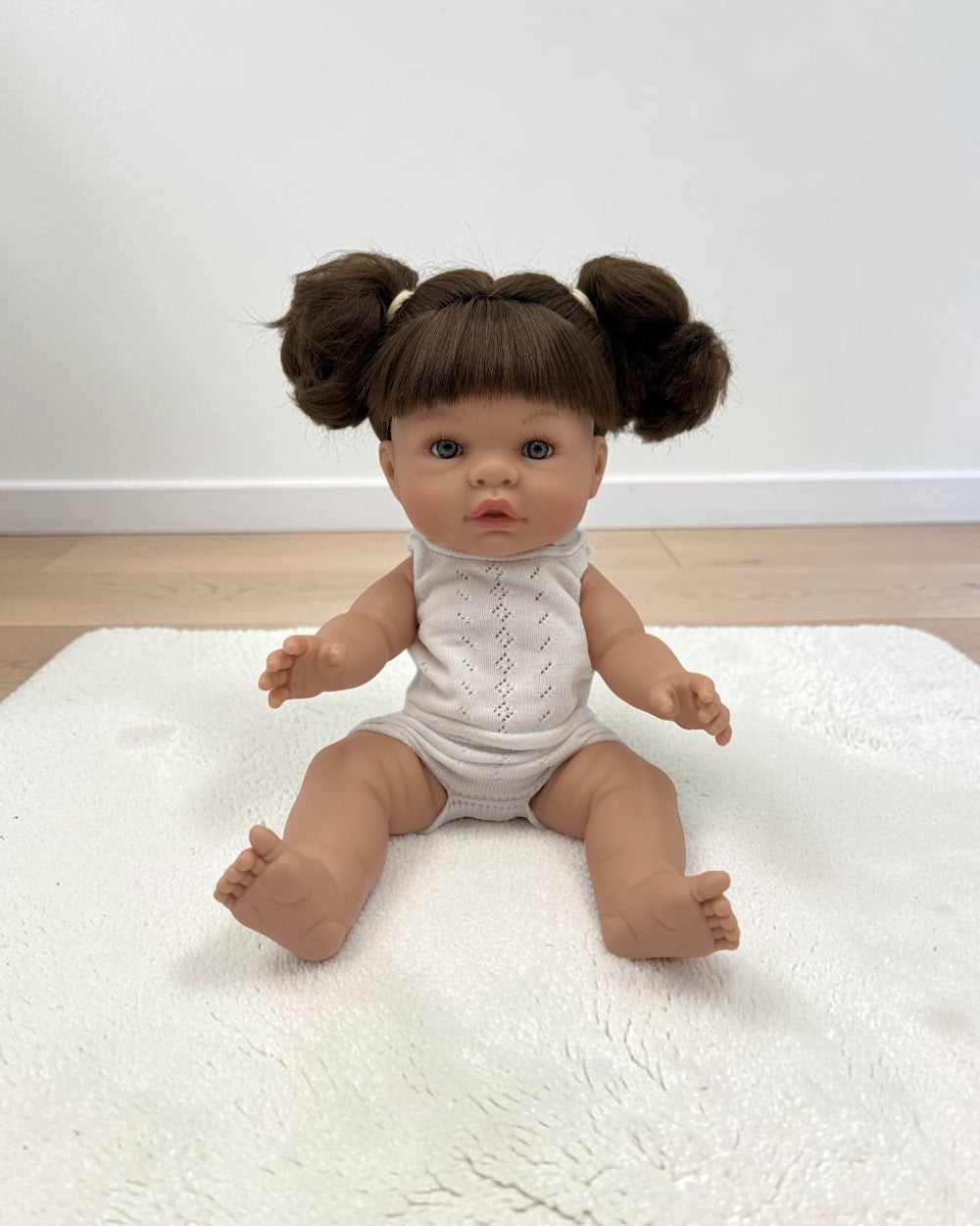 A realistic baby doll with brown hair in two buns sits on a white textured rug in a room with a white wall and light wood floor. The doll is wearing a white sleeveless knit bodysuit.