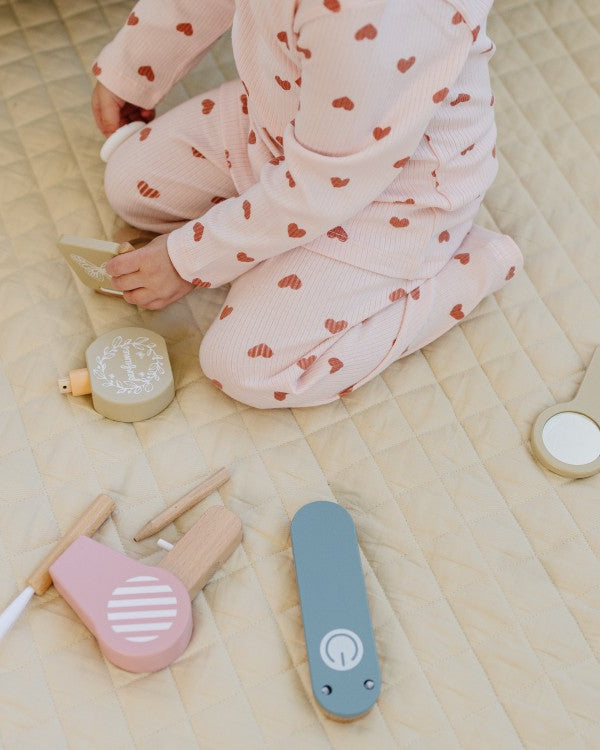 Child in pink pajama with heart patterns sitting on a bed surrounded by wooden toys.