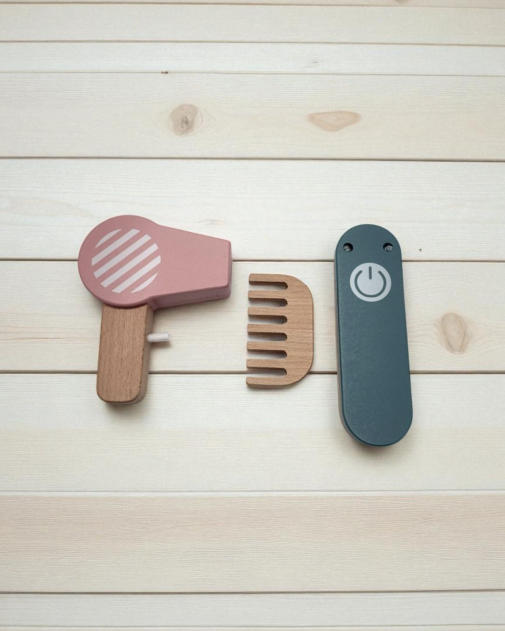 Set of wooden hair care tools including a pink hair dryer, brown comb, and blue straightener on a light wood background.