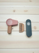 Set of wooden hair care tools including a pink hair dryer, brown comb, and blue straightener on a light wood background.