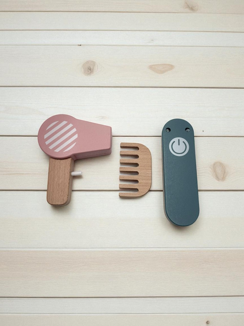 Set of wooden hair care tools including a pink hair dryer, brown comb, and blue straightener on a light wood background.