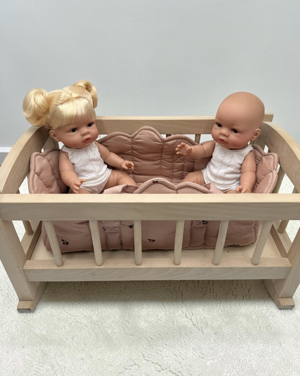 A miniature light wood doll crib with a pink quilted cushion, holding two baby dolls dressed in white outfits, set against a plain light gray wall and textured floor.