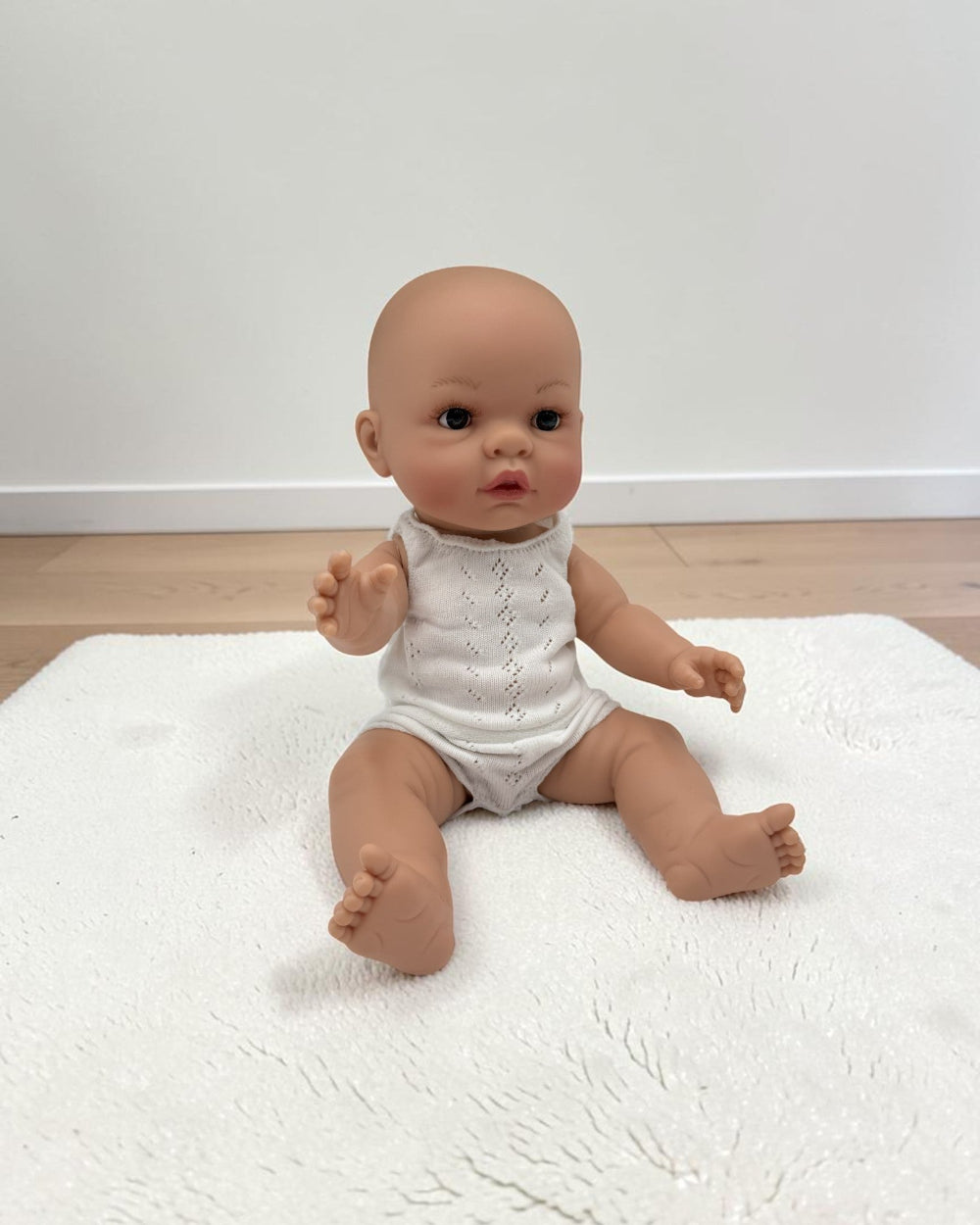 A baby doll with realistic skin and dark eyes sits on a white textured rug in a room with light wood flooring and a white wall.