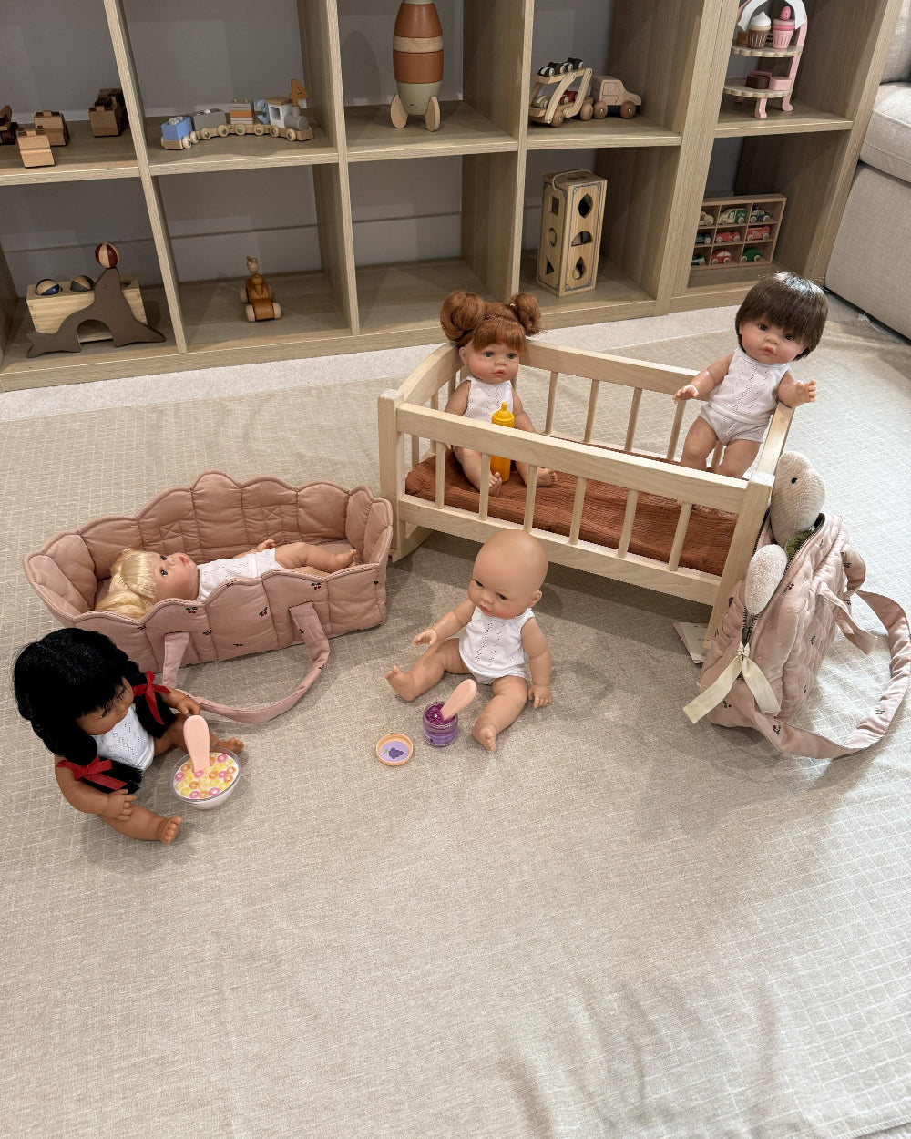 A doll play set arranged on a light beige fabric floor in front of a wooden toy storage shelf. The set includes a light wood baby crib with two dolls, a pink stroller with a doll lying inside, a pink bunny backpack, a rocking horse, and small toy accessories.