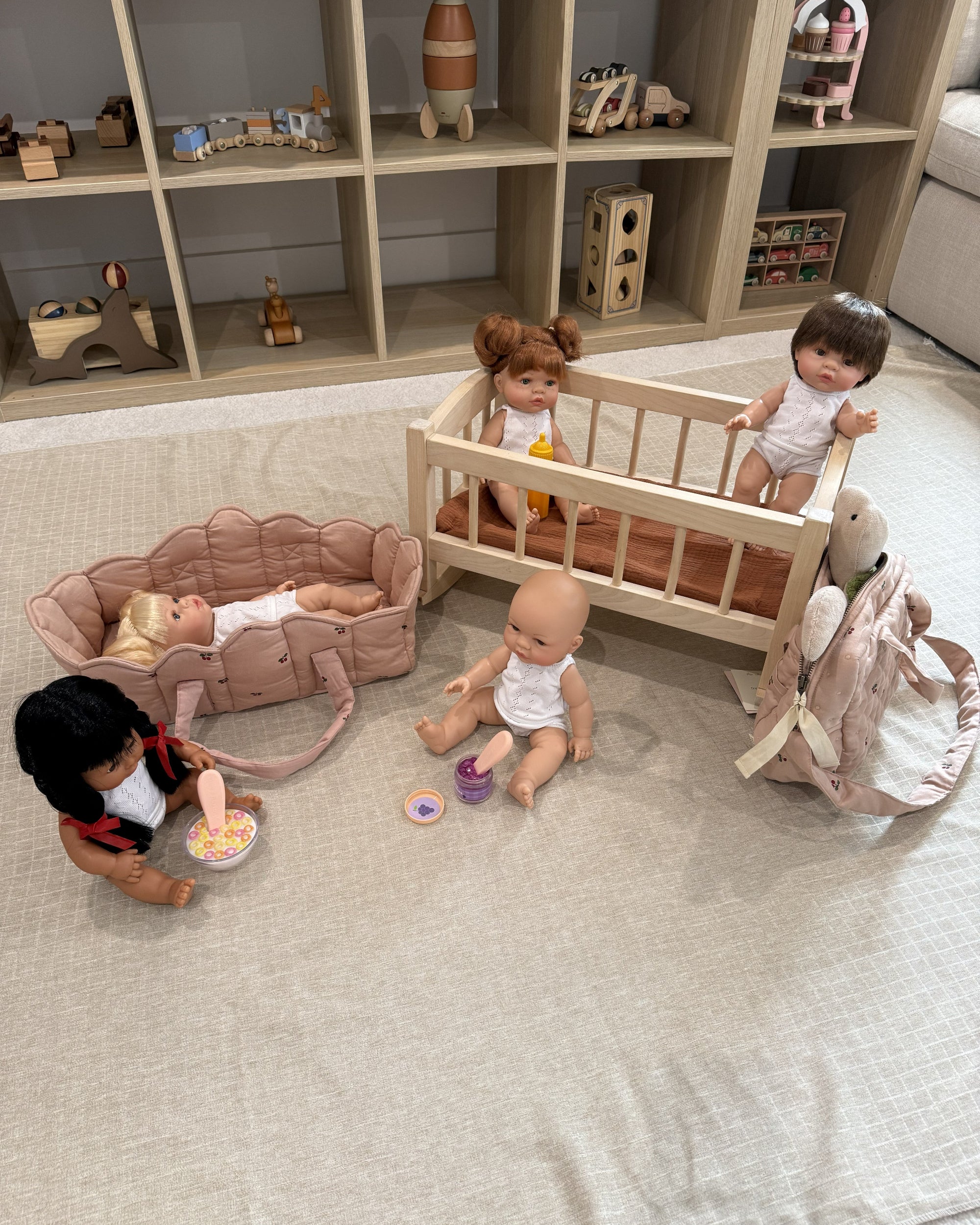 A doll play set arranged on a light beige fabric floor in front of a wooden toy storage shelf. The set includes a light wood baby crib with two dolls, a pink stroller with a doll lying inside, a pink bunny backpack, a rocking horse, and small toy accessories.