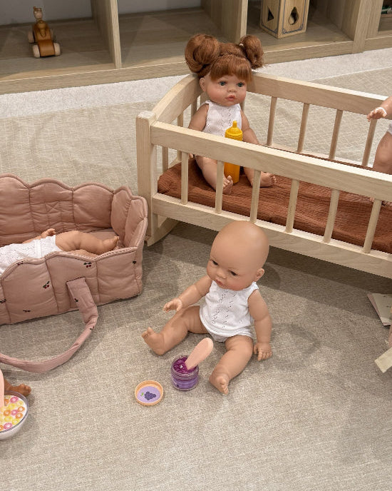A doll play set arranged on a light beige fabric floor in front of a wooden toy storage shelf. The set includes a light wood baby crib with two dolls, a pink stroller with a doll lying inside, a pink bunny backpack, a rocking horse, and small toy accessories.