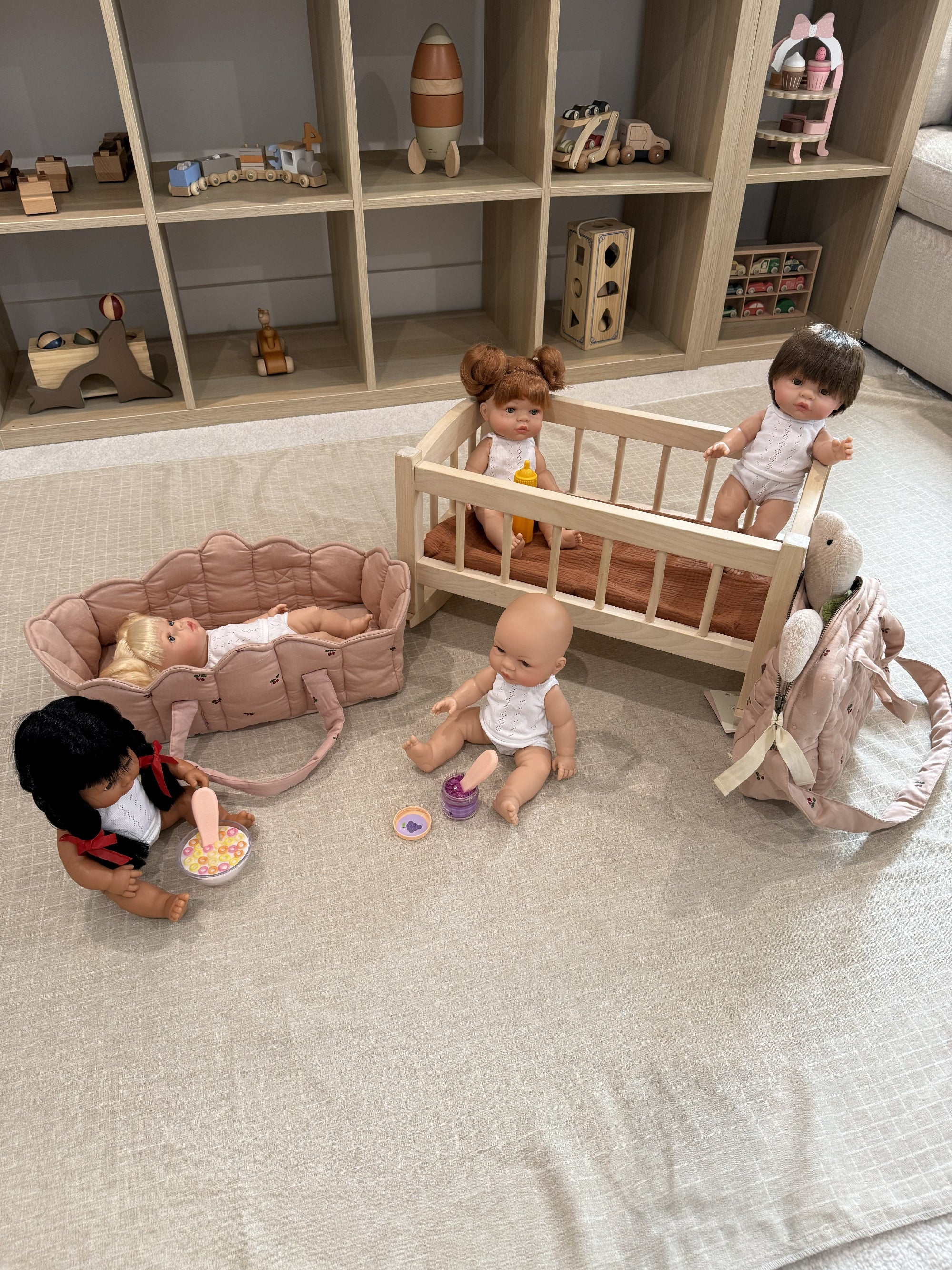 A doll play set arranged on a light beige fabric floor in front of a wooden toy storage shelf. The set includes a light wood baby crib with two dolls, a pink stroller with a doll lying inside, a pink bunny backpack, a rocking horse, and small toy accessories.