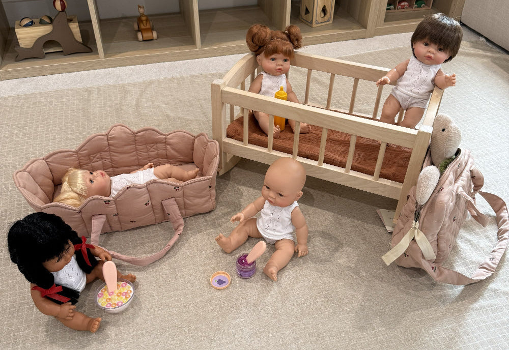 A doll play set arranged on a light beige fabric floor in front of a wooden toy storage shelf. The set includes a light wood baby crib with two dolls, a pink stroller with a doll lying inside, a pink bunny backpack, a rocking horse, and small toy accessories.