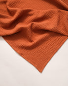 A cinnamon-colored muslin swaddle blanket laid out on a white surface.