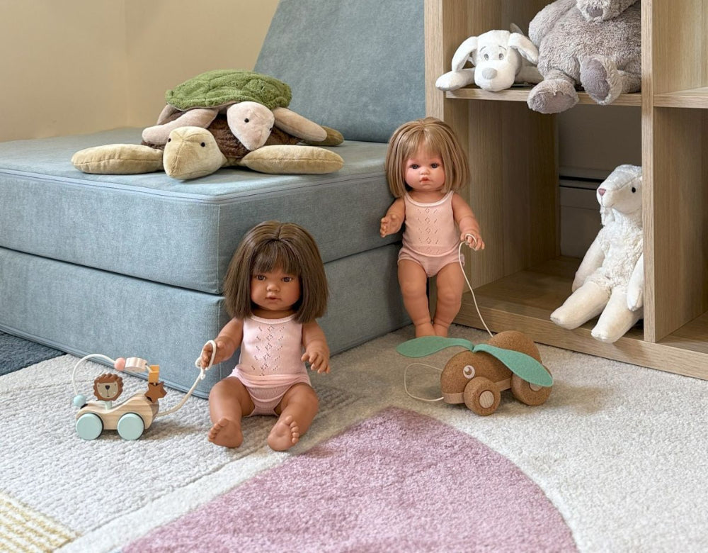 Two dolls playing with toys on a carpeted floor with a bookshelf in the background.