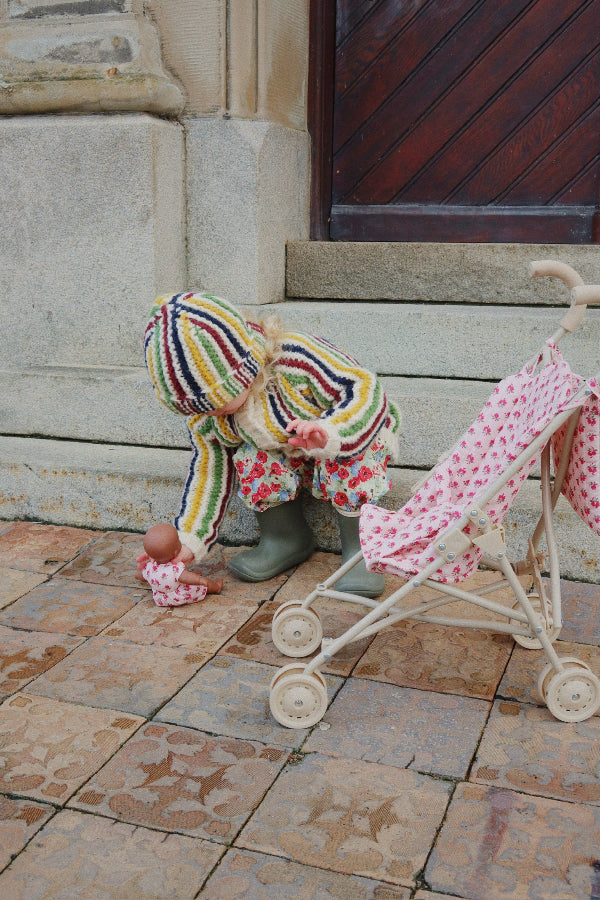 Child in colorful outfit playing with a baby doll and pink stroller on stone pavement.