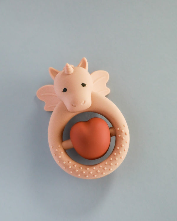 A silicone teether designed as a unicorn with a rose sand color heart-shaped element in the center, presented against a blue background.