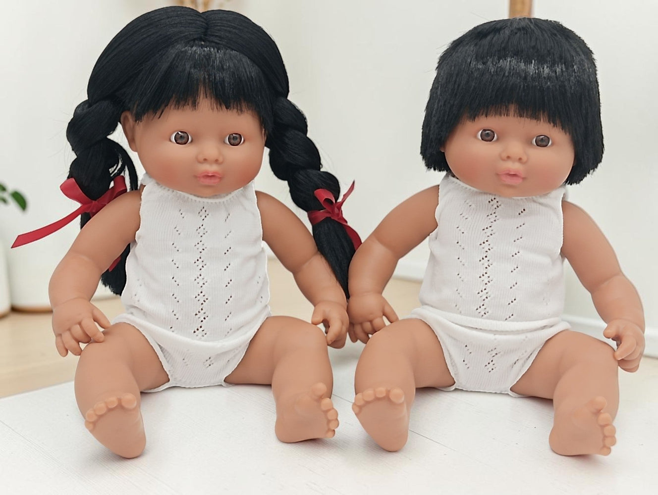 Two dolls with black hair and white outfits sitting on a light surface.