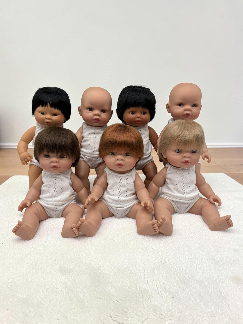Sevenrealistic baby dolls with various hair colors and skin tones, wearing white outfits, arranged on a white textured rug against a plain wall.