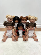 Seven realistic baby dolls with diverse hair colors and styles, wearing white outfits, arranged together on a white textured rug against a plain white wall.
