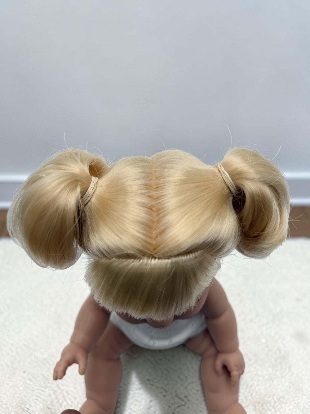 A doll viewed from behind, wearing a blonde hair accessory with two pigtails and a center part, seated on a white textured surface against a plain light gray wall.