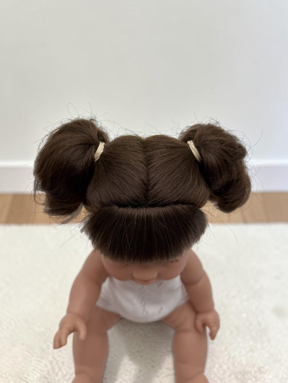 A doll with dark brown hair styled in two buns with small cream hair ties, wearing a white diaper, sitting on a white textured rug.