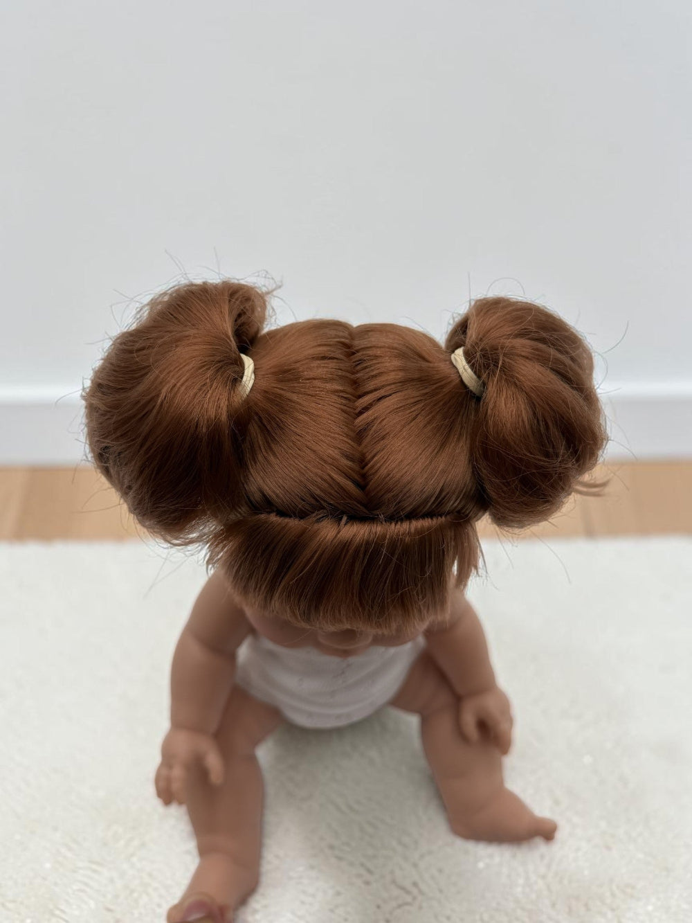 A close-up shot of a doll from behind, showing its detailed auburn hair styled in two buns with small cream elastic bands, wearing a white top, sitting on a white textured rug.