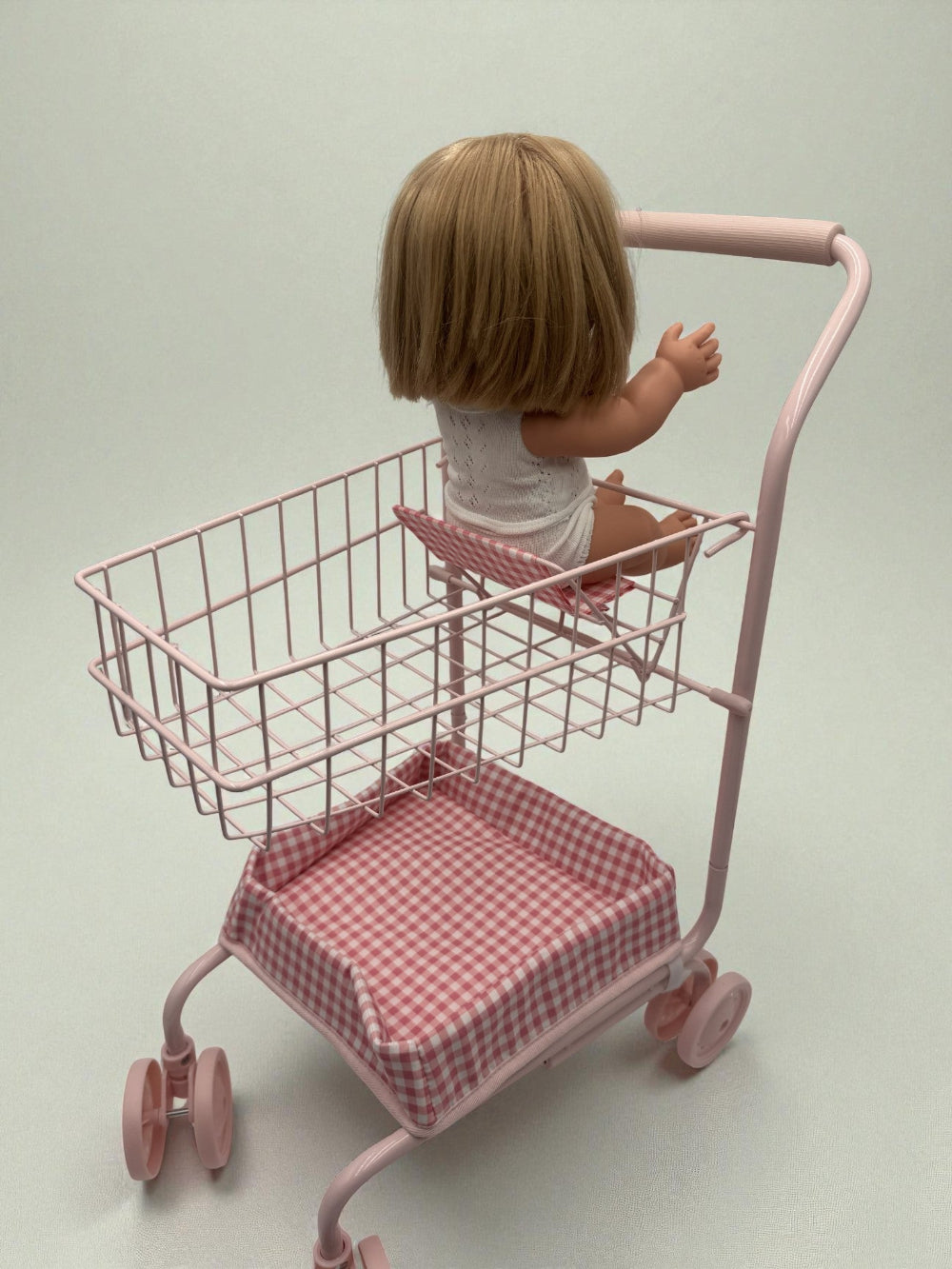 Doll sitting in a pink toy shopping cart with a checkered seat on a plain background
