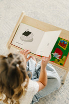A child sitting on a patterned carpet reads from an open wooden children's book stand. The book shows a simple house illustration, and colorful children's books are tucked into the side of the stand.