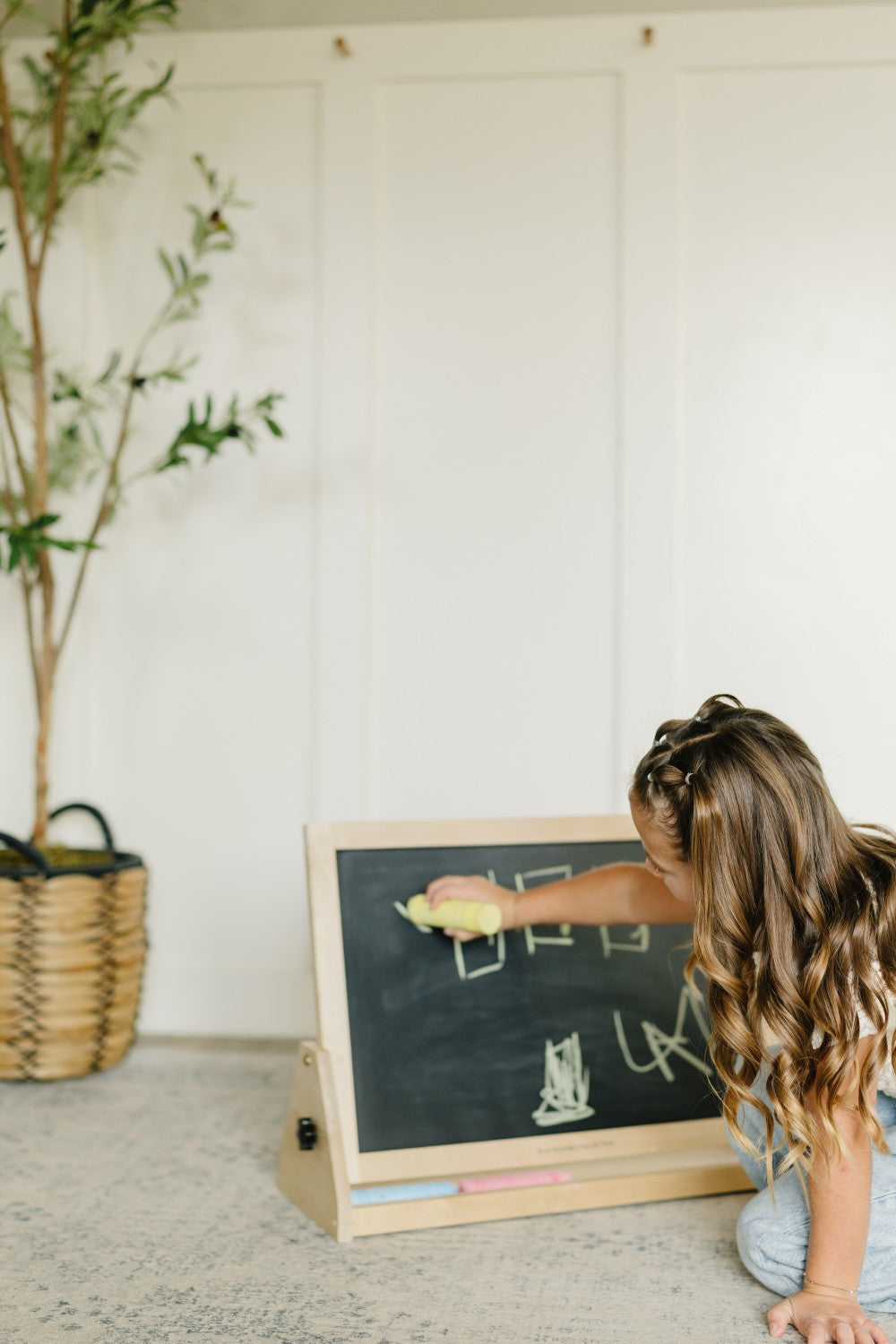 A young girl with long wavy brown hair kneels on a gray carpet, writing on a small wooden children's chalkboard. The chalkboard has a black slate face, a natural wood frame, and a light wood base with chalk sticks.
