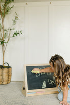 A young girl with long wavy brown hair kneels on a gray carpet, writing on a small wooden children's chalkboard. The chalkboard has a black slate face, a natural wood frame, and a light wood base with chalk sticks.