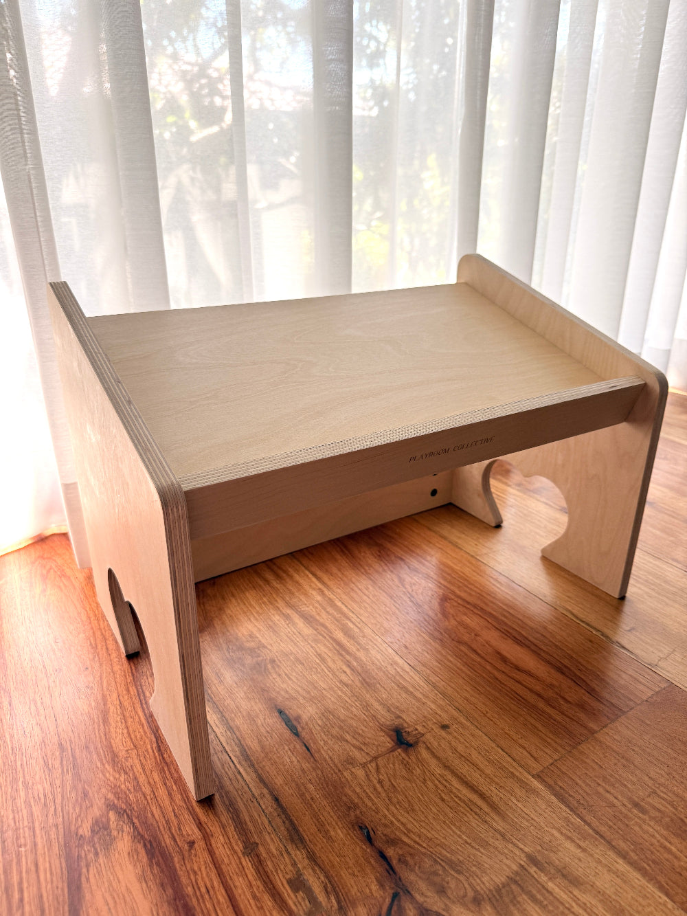 A light natural wood folding desk or stand with a slanted top, shown on a hardwood floor in front of sheer white curtains.