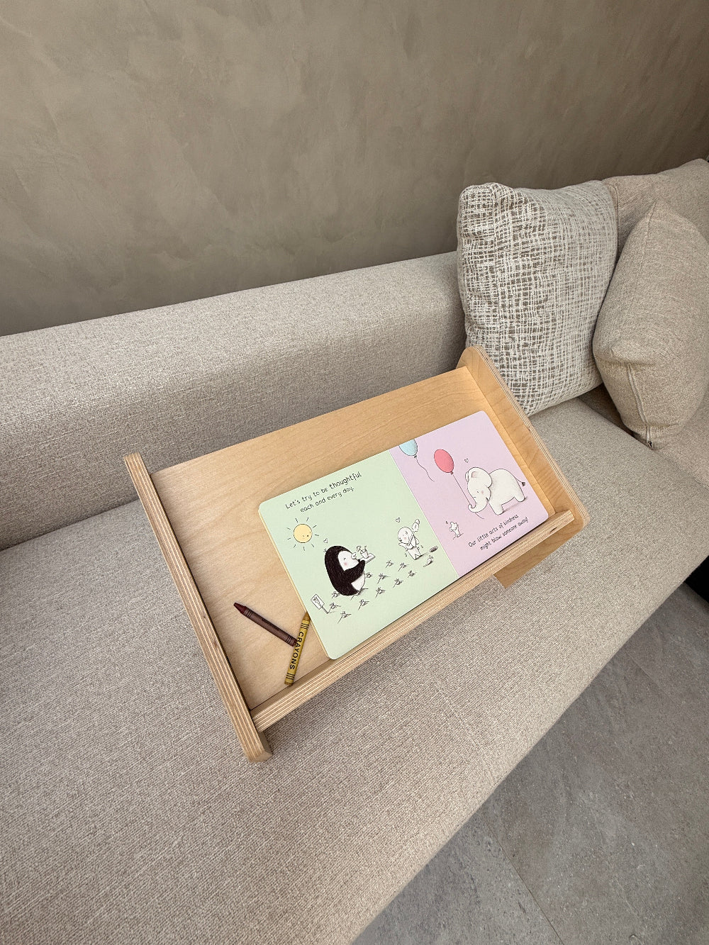 A light wood lap desk tray resting on a gray fabric sofa, holding a pastel cartoon card and a pencil.