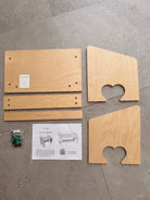 Wooden furniture assembly kit parts laid out on a gray tiled floor, including two curved top panels, two rectangular side panels, a long narrow piece, a printed instruction sheet, and a small bag of screws and hardware.