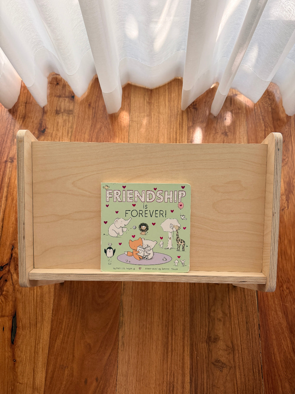 A light natural wood toy shelf or bookcase placed on a hardwood floor in front of white sheer curtains. A square illustrated book or card titled "Friendship is Forever!" rests on the shelf, showing pastel artwork with animals and hearts.