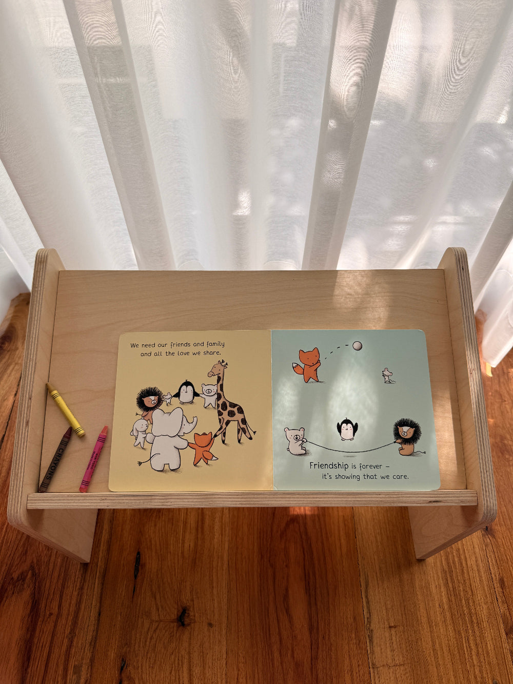 A small wooden children's book stand on a wooden floor in front of sheer white curtains. The stand holds an open children's book with two illustrated pages and two small colored pencils.